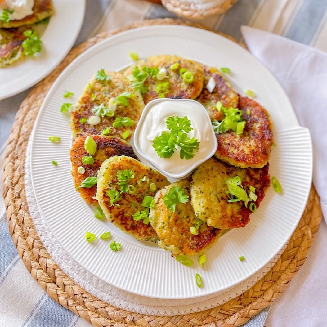 Belarusian draniki potato pancakes arranged around a small bowl of sour cream on a white plate, topped with chopped spring onion and parsley on a woven mat.