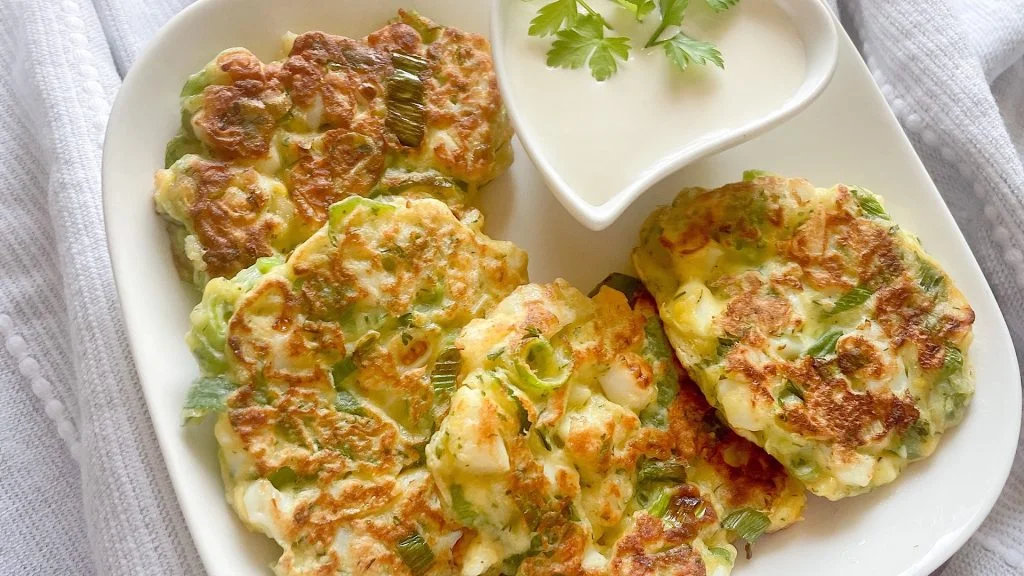 Overhead view of crispy egg and spring onion fritters on a square white plate beside a heart-shaped bowl of sour cream garnished with parsley, set on a wooden board and linen cloth.
