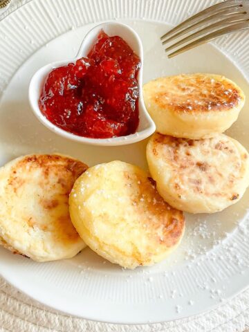 Easy Ricotta Syrniki Recipe plated with powdered sugar and a side of red berry jam.