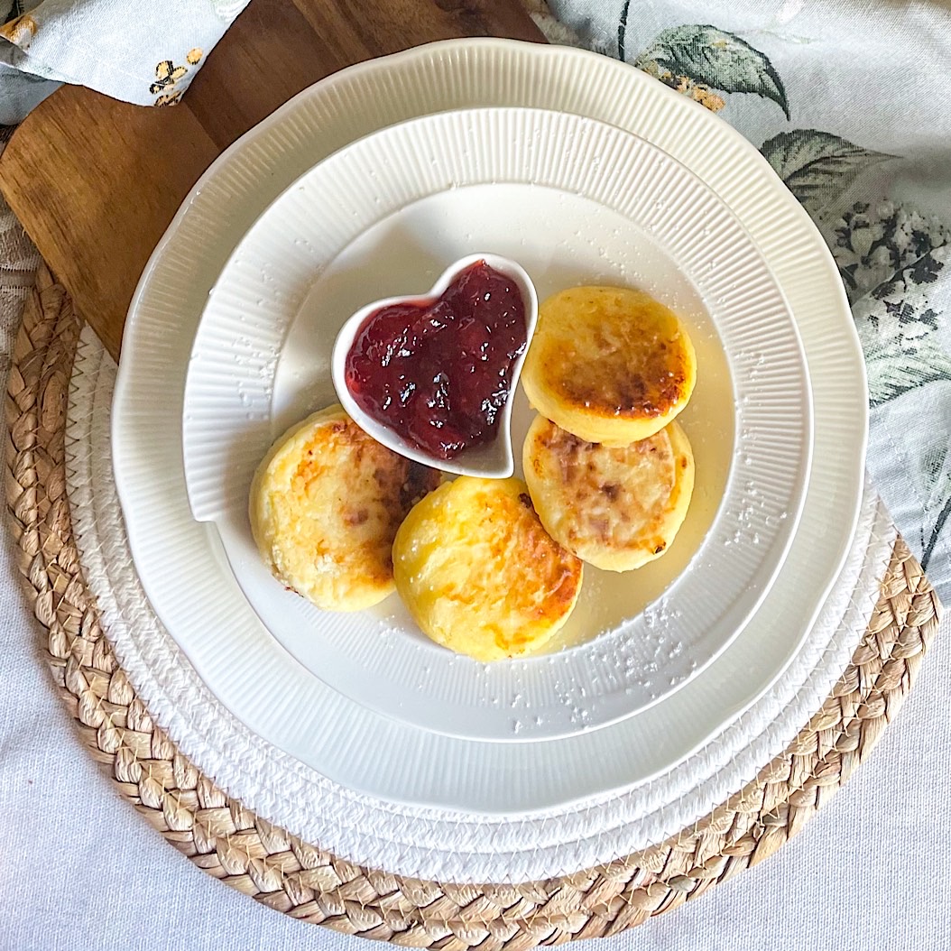 Golden Ricotta Syrniki served on a white plate with strawberry jam in a heart-shaped bowl.