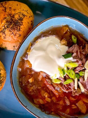 Borscht on meaty bones served in a blue bowl with sour cream, spring onions, sliced garlic, and baked buns on a wooden board