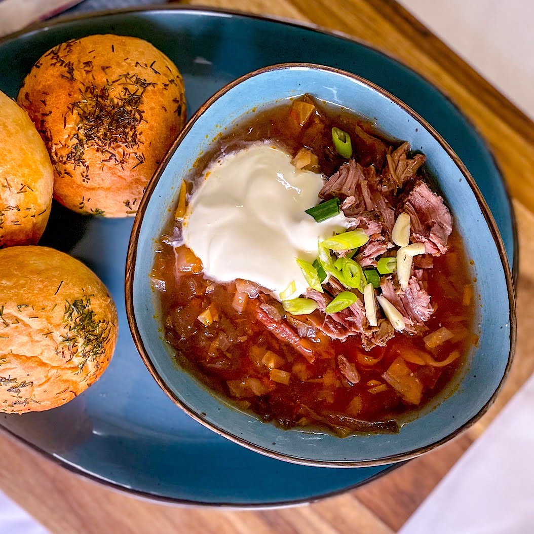 Bowl of borscht on meaty bones stock with shredded beef, sour cream, and fresh spring onions, served with baked dill buns