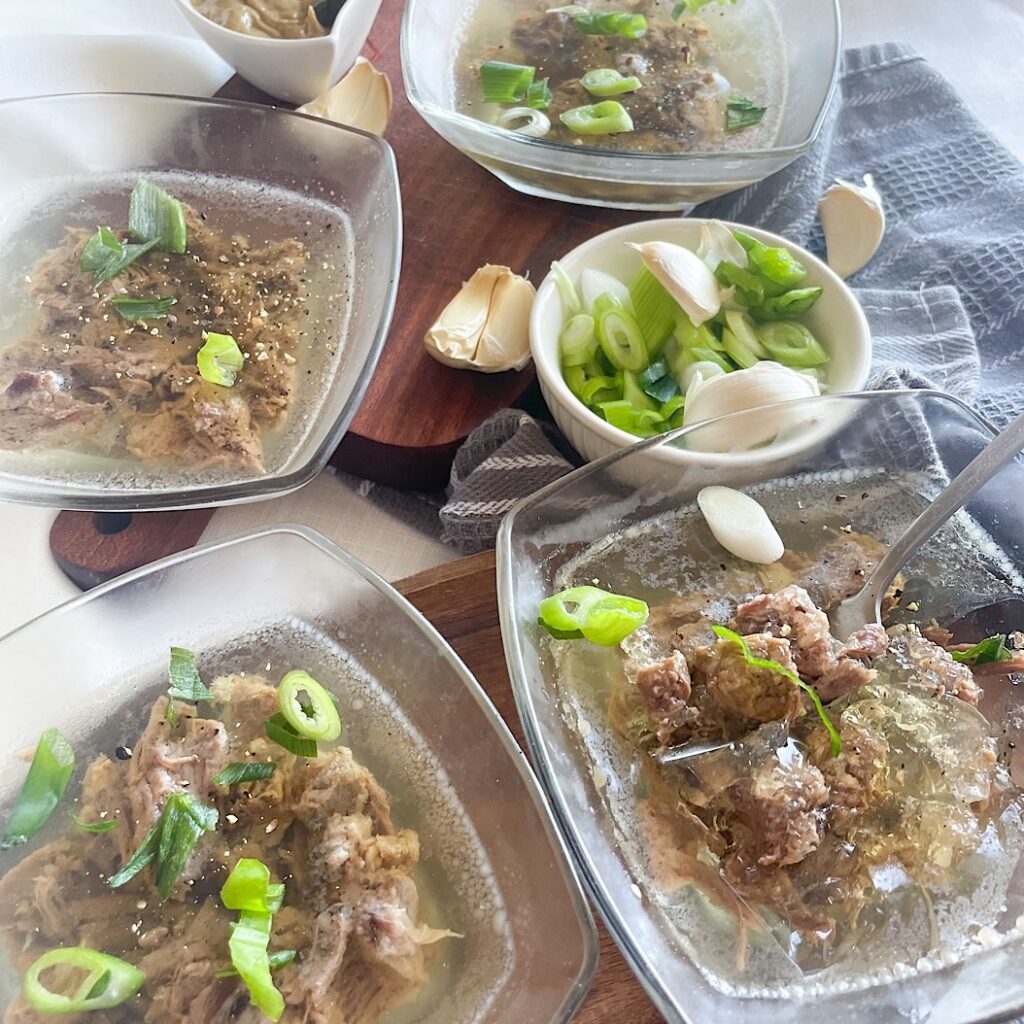 : An overhead view of a traditional spread featuring multiple bowls of Beef Kholodets (Beef Aspic) topped with green onions, served alongside fresh garlic cloves and a small dish of horseradish.
