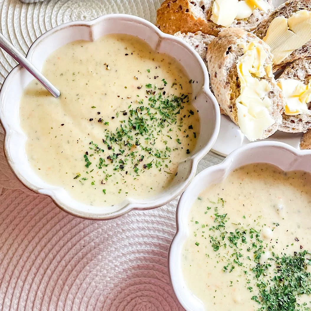 Creamy vegetable soup in a scalloped white bowl topped with chopped parsley and black pepper, buttered bread behind