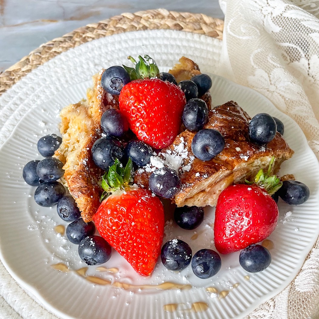 French toast croissant casserole topped with fresh strawberries, blueberries, and a dusting of icing sugar on a white plate.