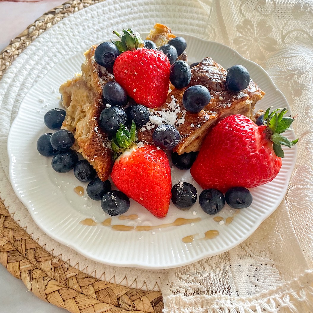 French toast croissant casserole topped with fresh strawberries, blueberries, and a dusting of icing sugar on a white plate.