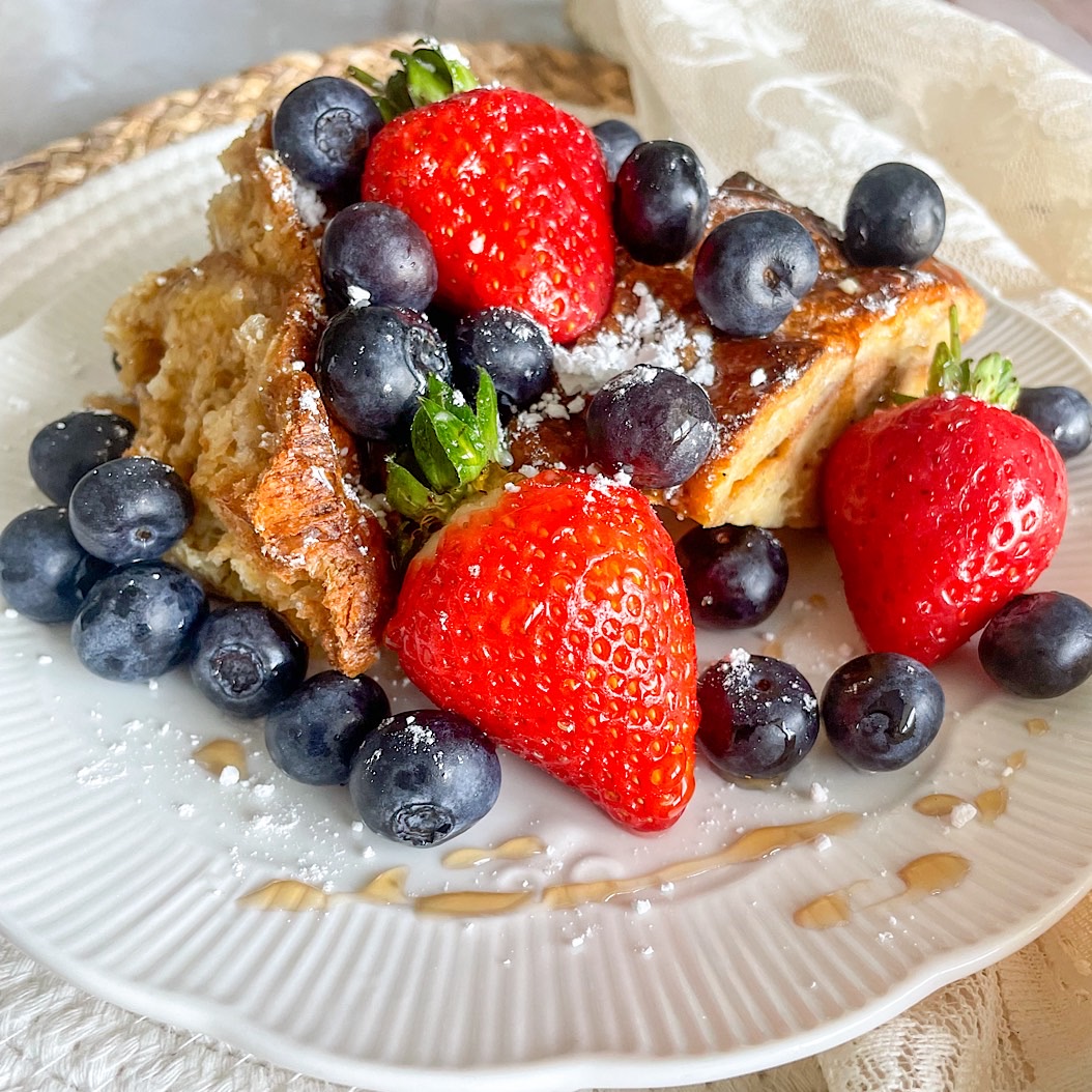 French toast croissant casserole topped with fresh strawberries, blueberries, and a dusting of icing sugar on a white plate.