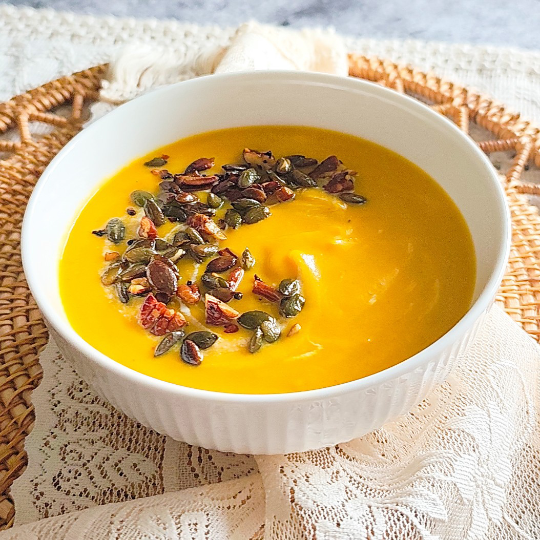 Bowl of creamy butternut squash soup topped with pecans and pumpkin seeds on lace tablecloth