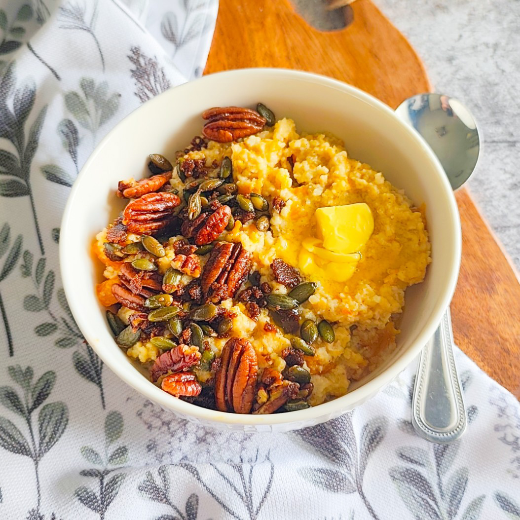 Bowl of pumpkin millet porridge topped with pecans, pumpkin seeds, and melting butter on a wooden board