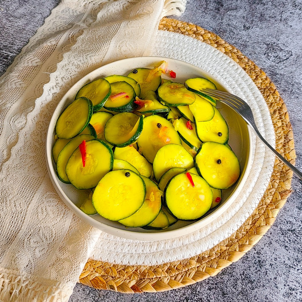 Overhead view of pickled courgettes in a patterned bowl on a rustic woven tablecloth.