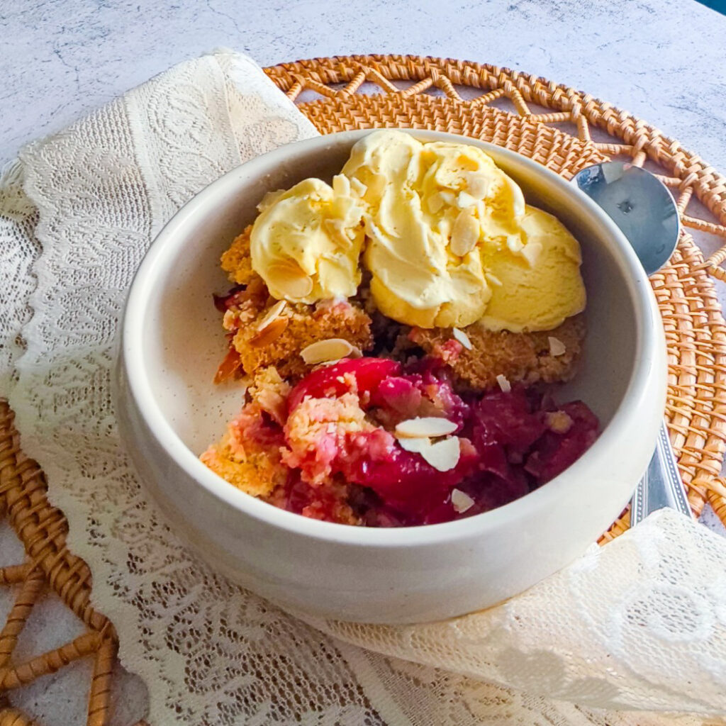 Bowl of plum crumble topped with vanilla ice cream and almond flakes on a lace cloth.