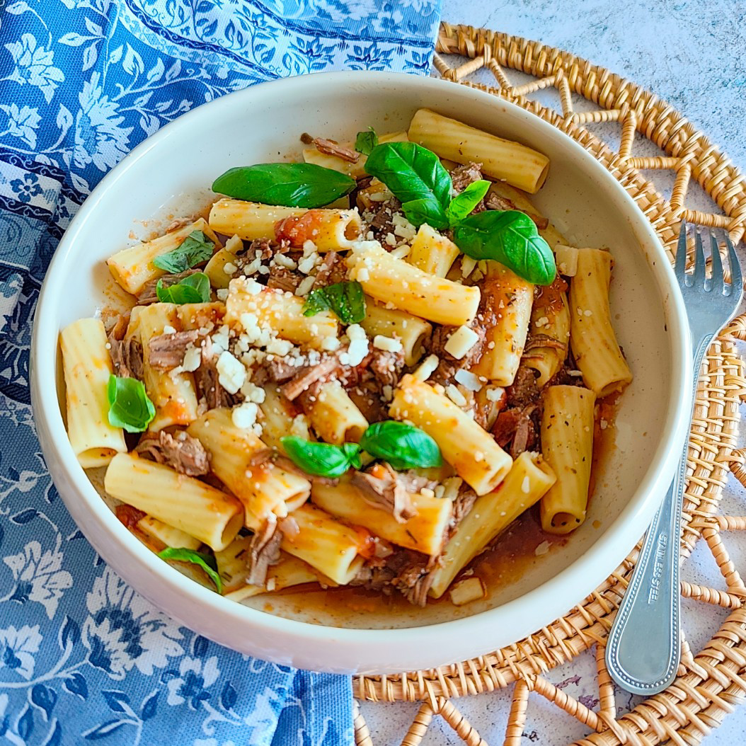 Bowl of slow cooked beef pasta with rigatoni, shredded beef, Parmesan, and fresh basil on a woven placemat.
