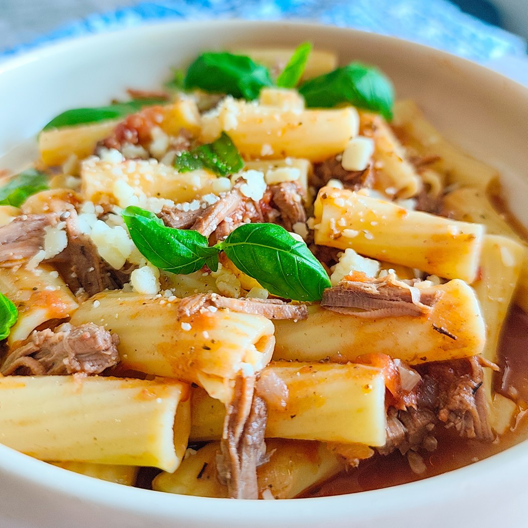 Close-up of rigatoni pasta with shredded slow cooked beef, tomato sauce, basil leaves, and grated cheese.
