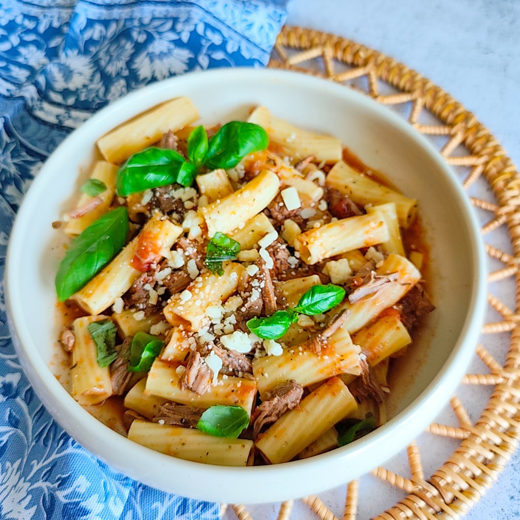 Rustic slow cooked beef pasta served in a white bowl with Parmesan and basil, placed on a blue floral napkin.