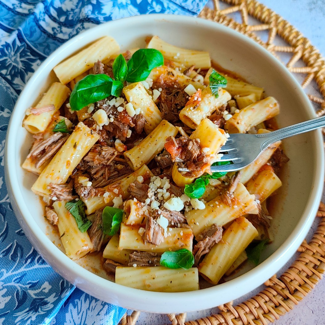 Fork lifting a bite of slow cooked beef pasta with rigatoni, tomato sauce, and fresh basil from a rustic bowl.