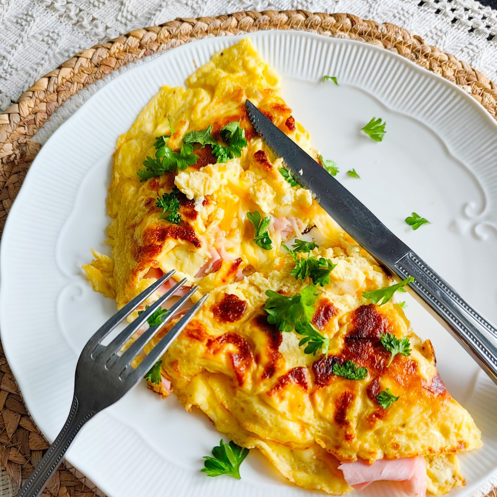 Ham and cheese omelette served on a white plate, topped with parsley and ready to eat with fork and knife.