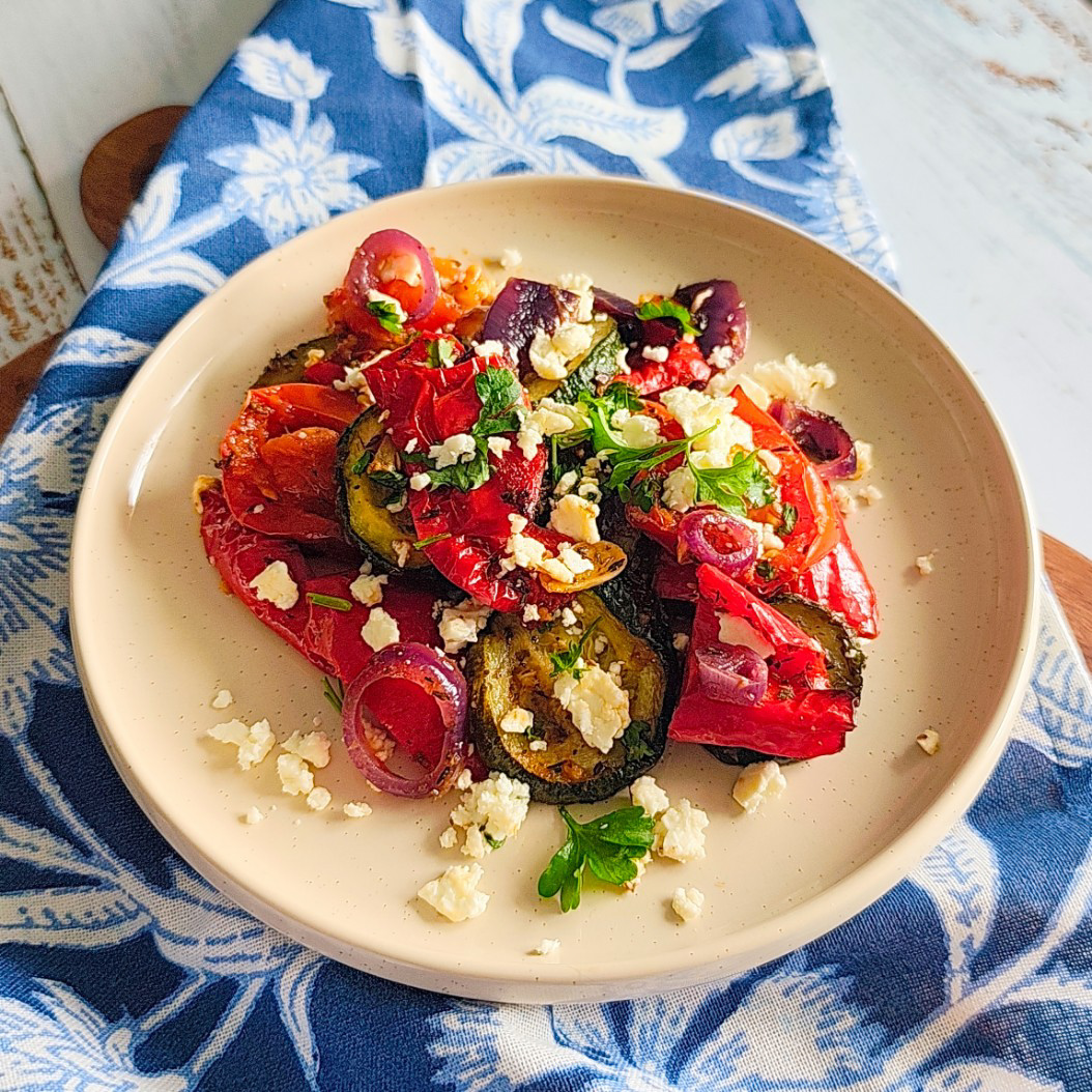 Roasted courgettes, red peppers, onions, and tomatoes with feta on a beige plate and wooden board.