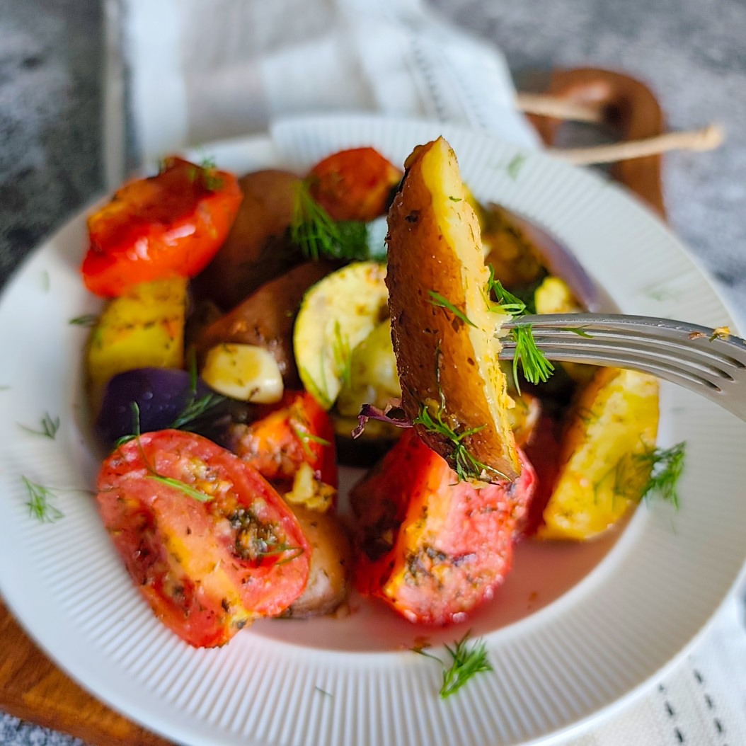 Fork lifting a wedge of potato from a bowl of colourful roasted vegetables with herbs.