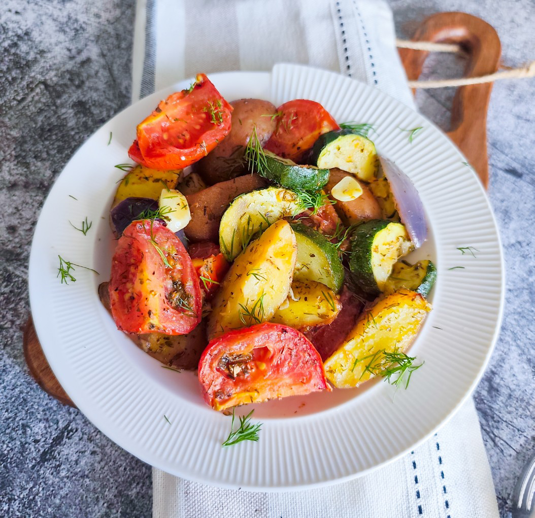 Overhead view of roasted vegetables garnished with dill in a white bowl on a wooden board.