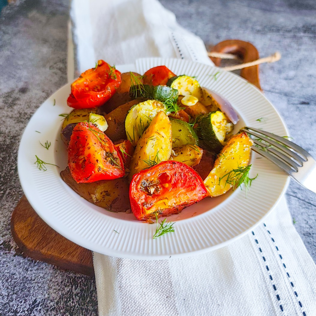 Plate of roasted vegetables with potatoes, courgettes, tomatoes, and fresh dill on a white dish.