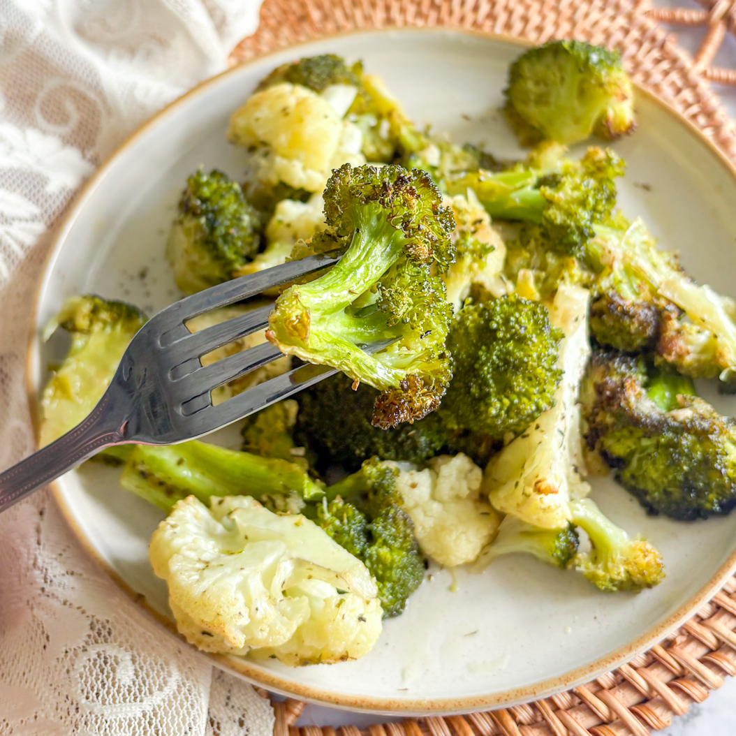 Fork holding a floret of roasted broccoli over a plate of roasted broccoli and cauliflower.