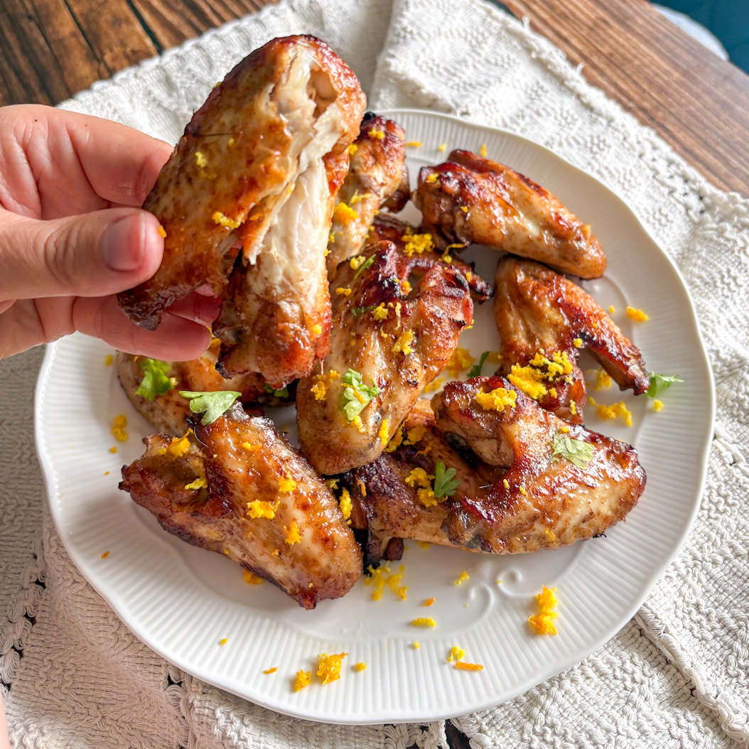 Hand holding a Five Spice Chicken Wing with juicy interior, plate of wings in background with zest and herbs.