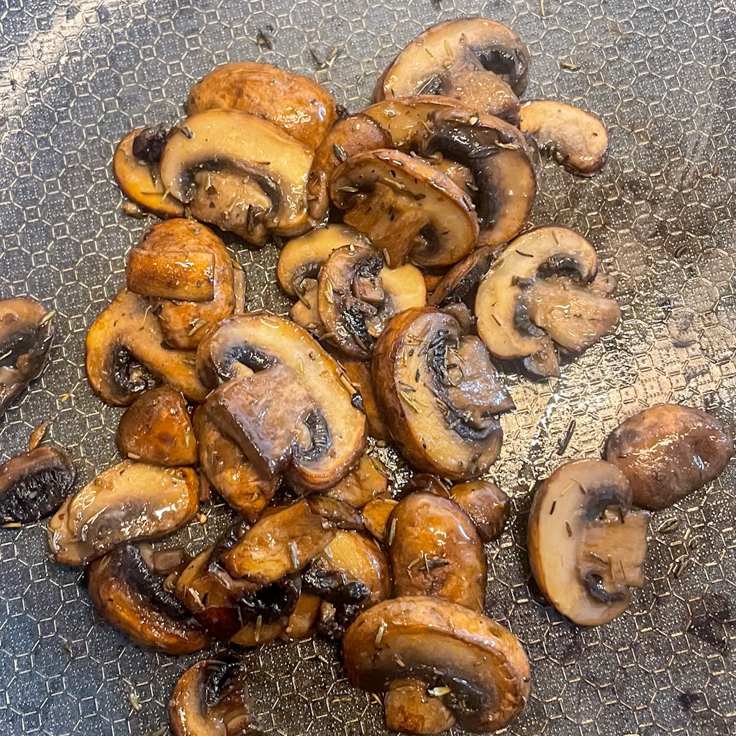 Sliced mushrooms frying in a skillet with butter and thyme for mushroom omelette filling.