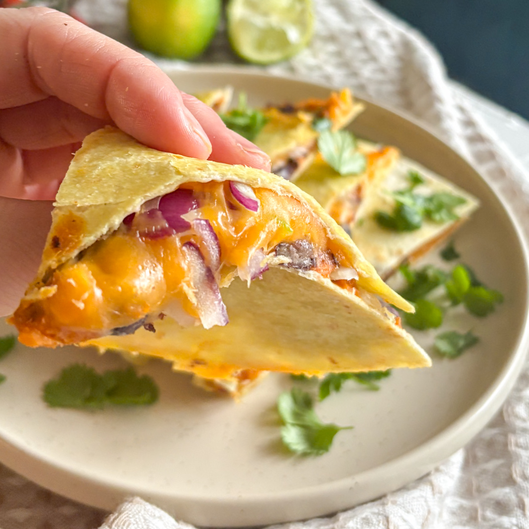 A hand holding a wedge of roasted sweet potatoes and black beans quesadillas to show the melted cheese and filling of this delicious recipe.