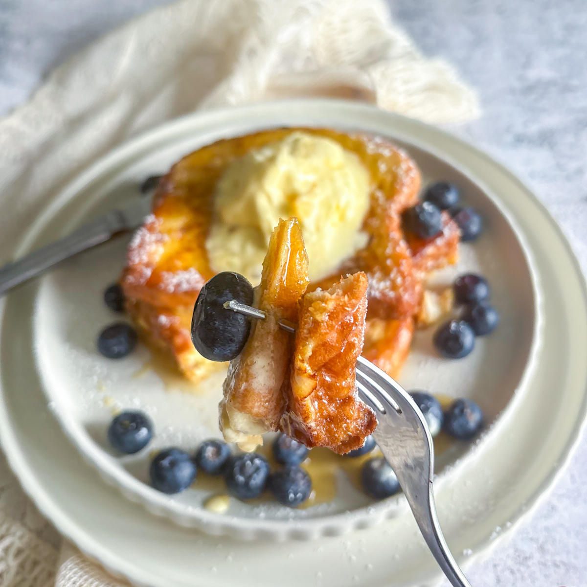 A close-up of a fluffy, sweet bite of Brioche French Toast on a fork with a fresh blueberry and syrup, showing the soft interior and crisp edge.