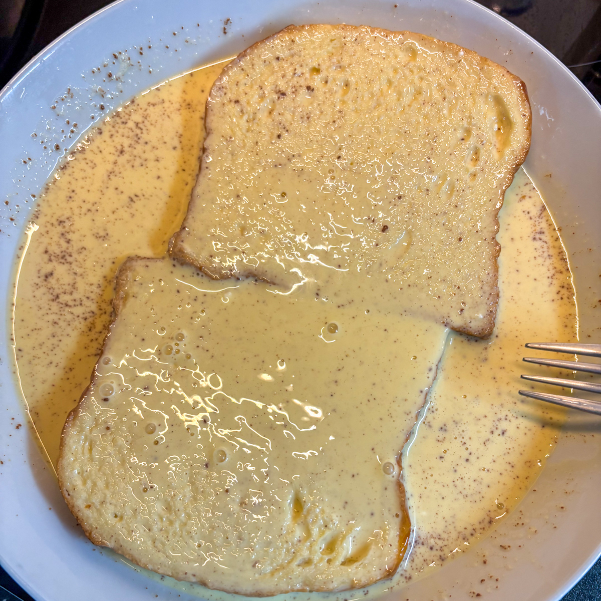 Thick slices of the best stale bread for French toast soaking in a rich egg custard inside a shallow bowl.
