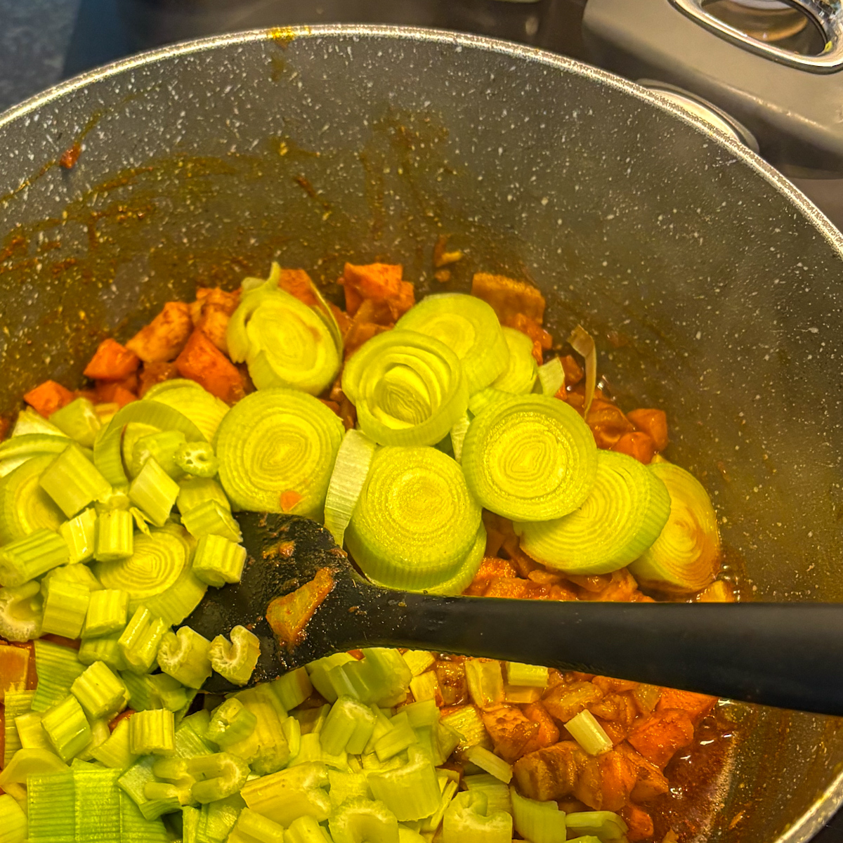 Adding chopped leek rounds and celery ribs to the spiced root vegetable mixture in the heavy-bottomed pot.