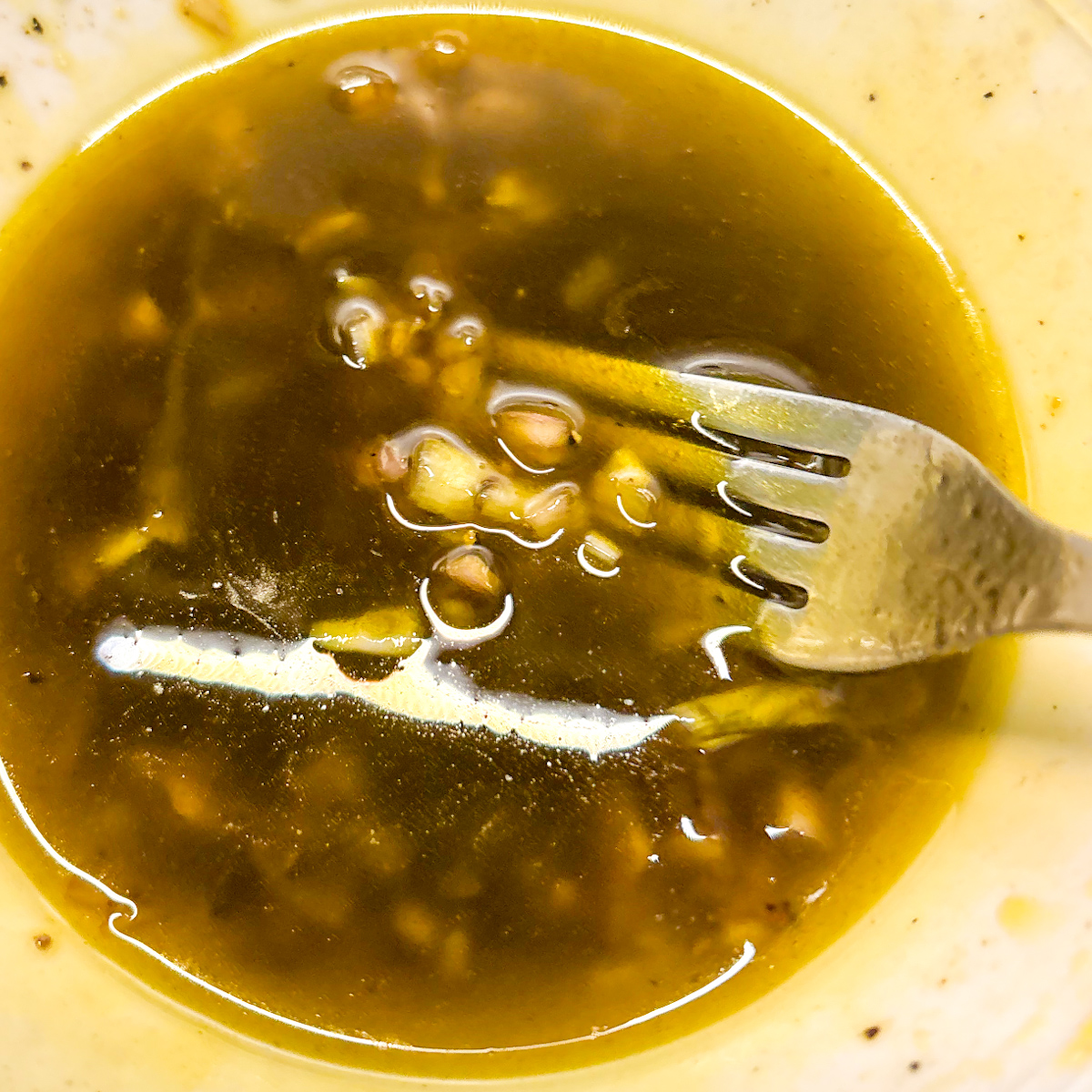 A close-up photograph of a fork whisking a tangy balsamic vinaigrette dressing for a crunchy chickpea salad in a small glass bowl, showing the emulsification.