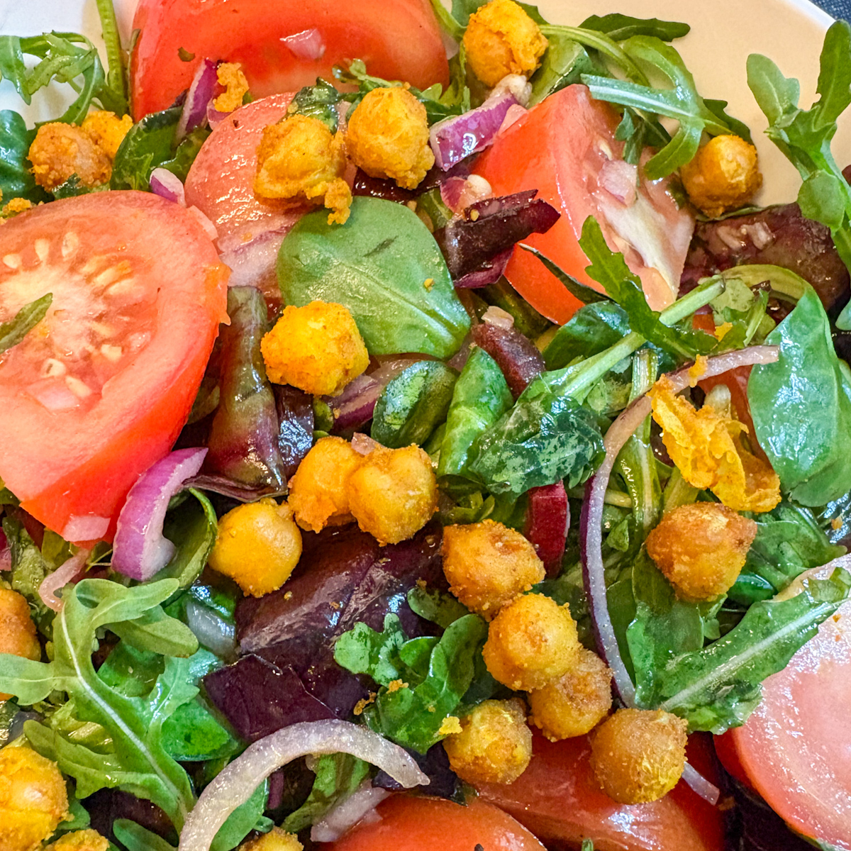 A top-down photograph showing bare hands tossing fresh mixed greens, red tomato wedges, and sliced red onion in a large ceramic bowl to prepare a leafy chickpea salad.