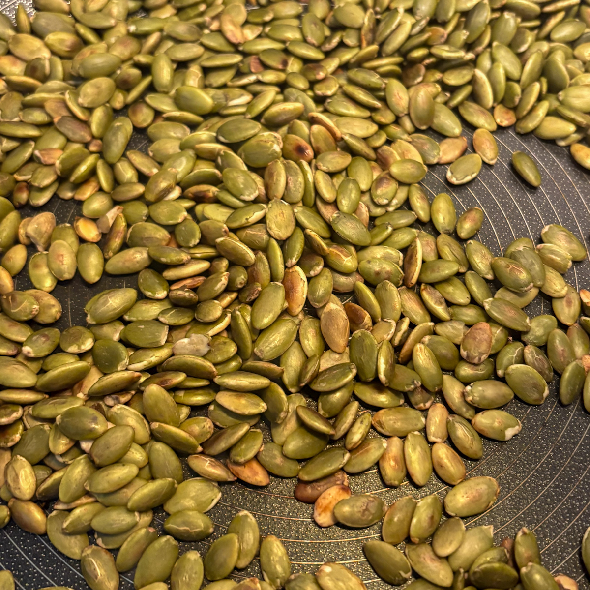 Raw green pumpkin seeds being dry-toasted in a dark, textured frying pan.