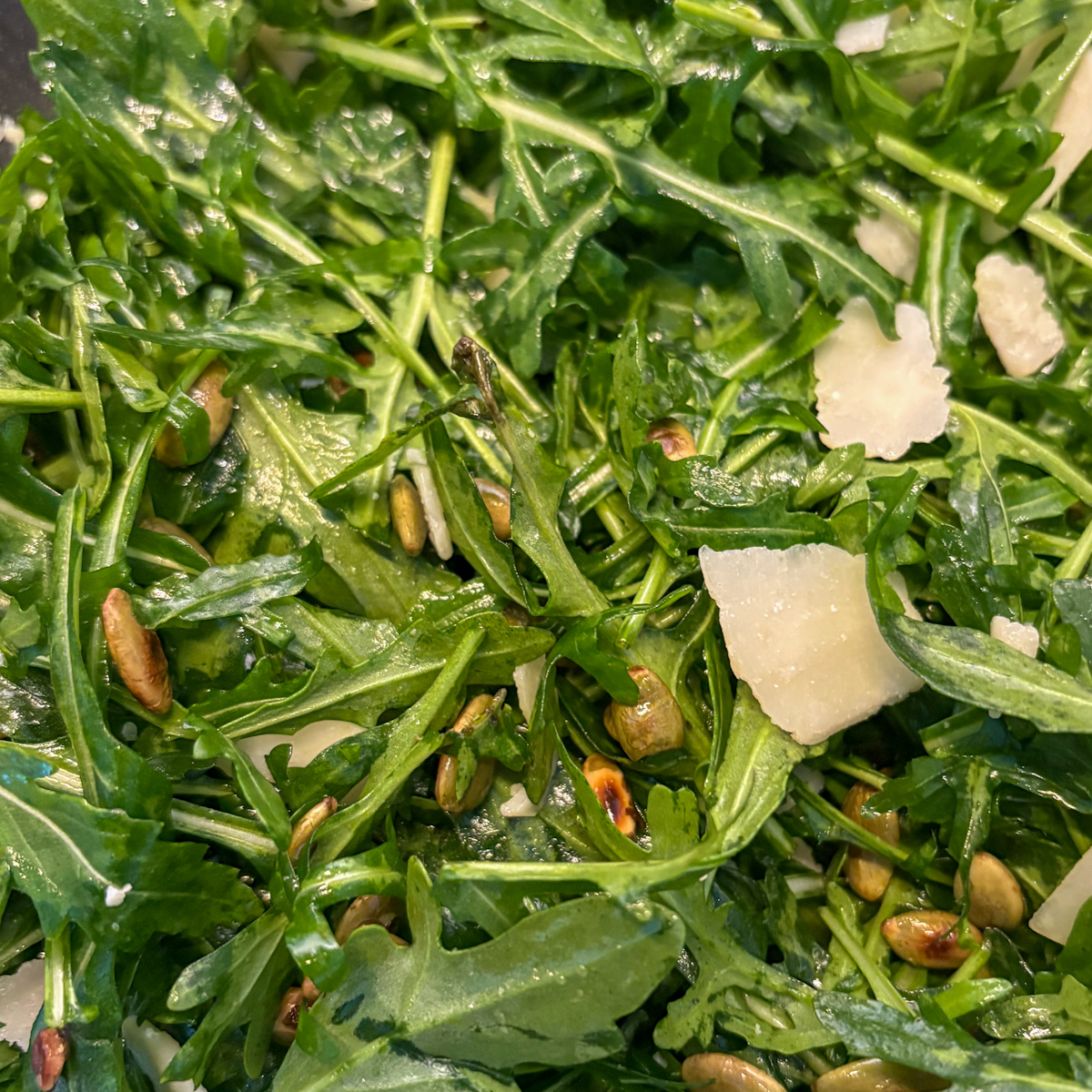 Close-up texture of fresh rocket leaves lightly coated in a tangy vinaigrette, mixed with toasted pumpkin seeds and parmesan pieces.