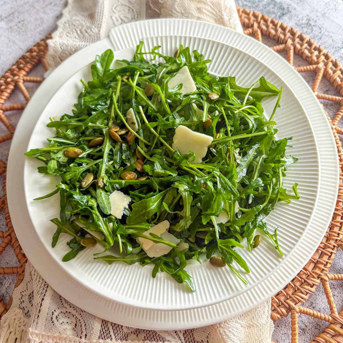 Top-down view of an easy rocket salad topped with parmesan shavings and toasted pumpkin seeds, served on a white fluted plate over a woven placemat.