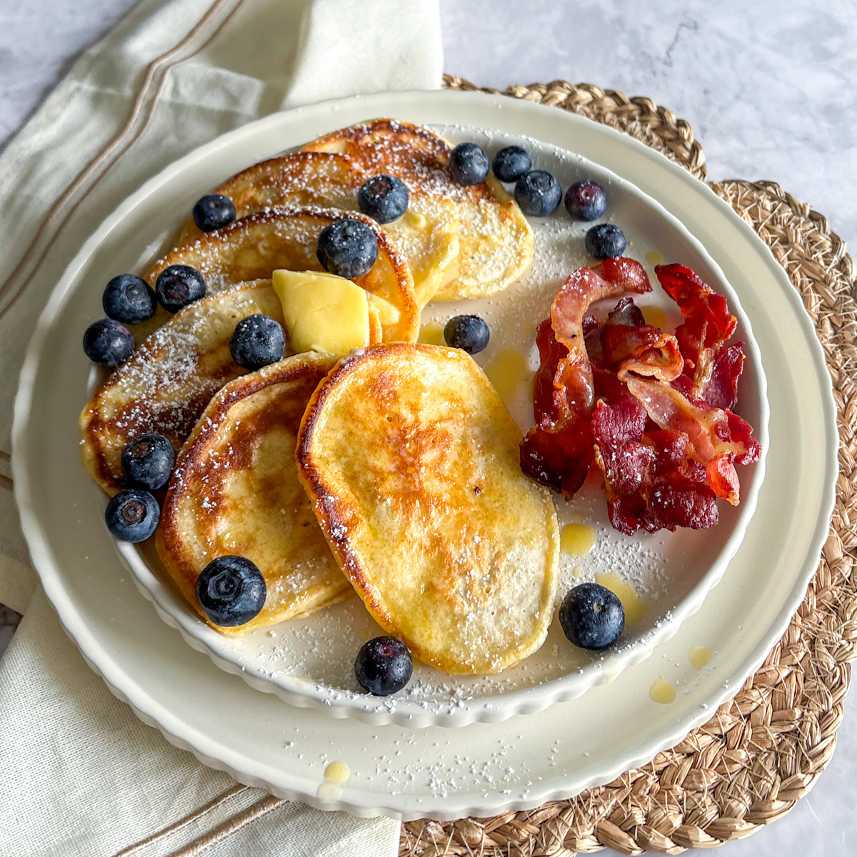 Top-down view of a thick, fluffy pancake stack being drizzled with maple syrup alongside a side of savory roasted bacon.