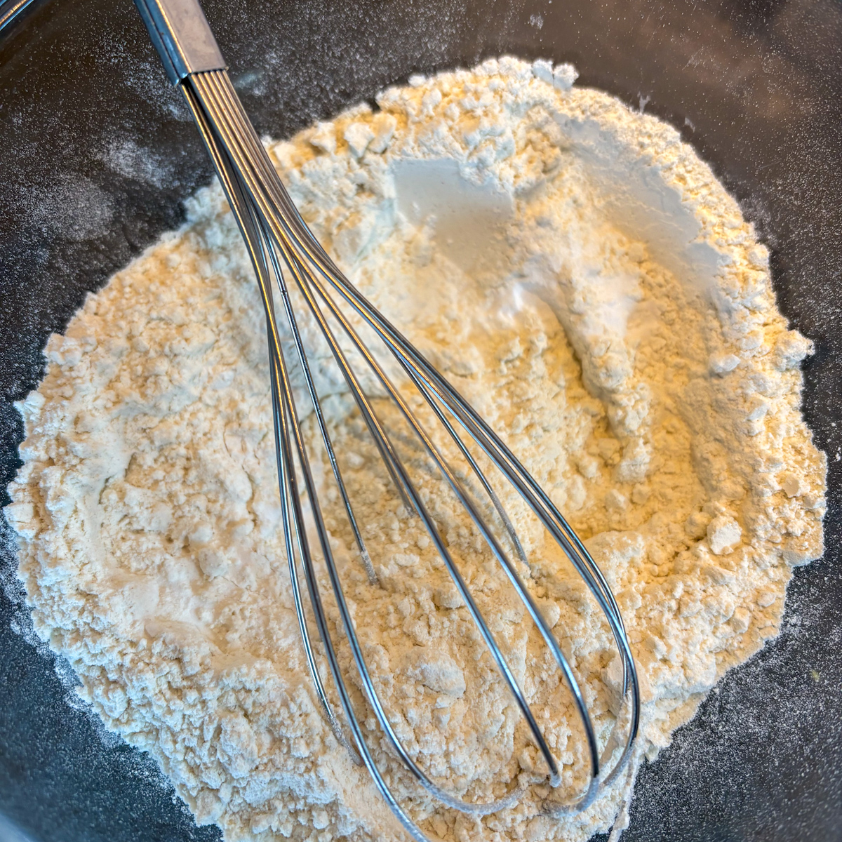 A glass bowl with a whisk mixing the plain white flour, sugar, and raising agents dry mixture for Homemade Buttermilk Pancakes.
