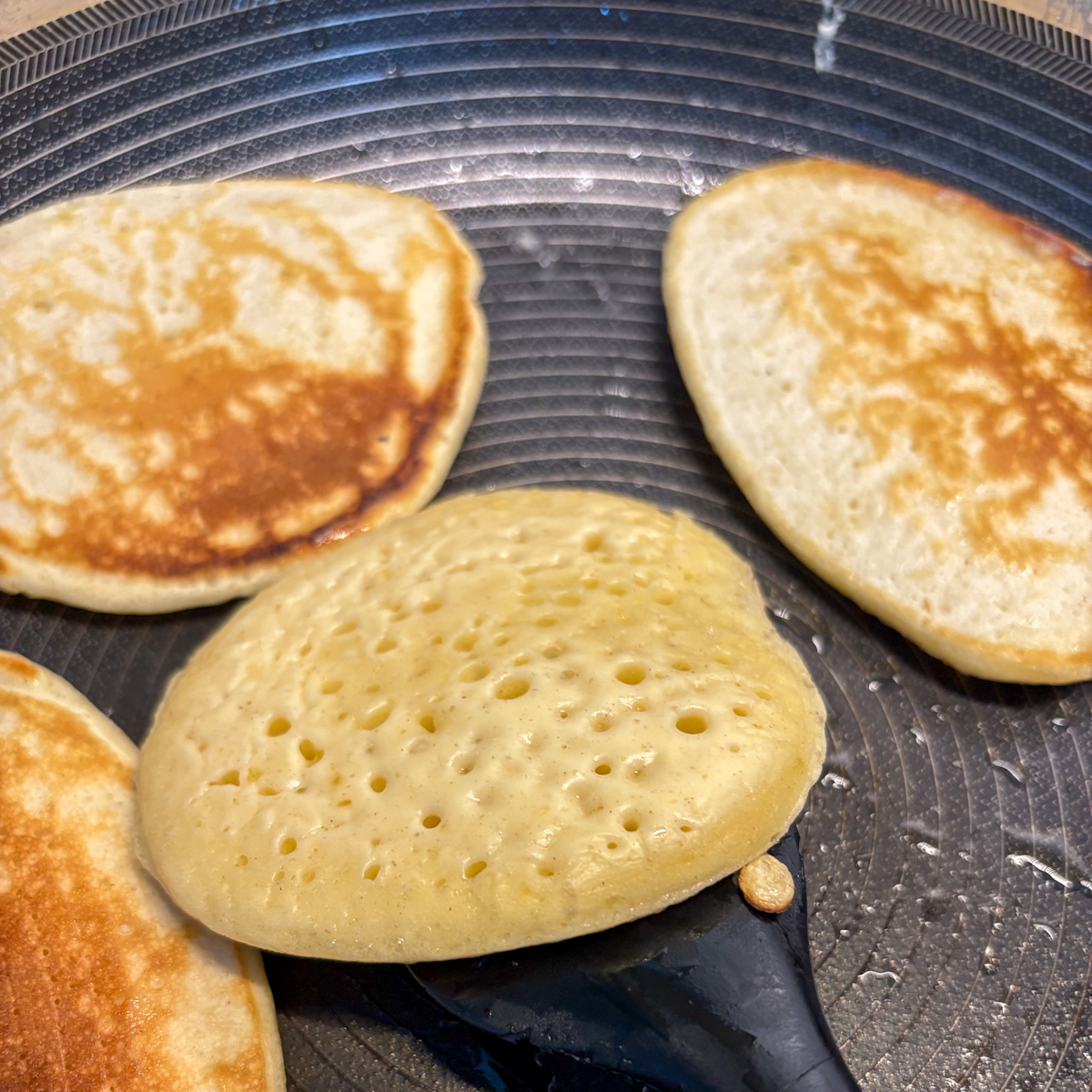 A golden-brown, thick buttermilk pancake cooking in a frying pan on medium-high heat with small bubbles forming on the surface.
