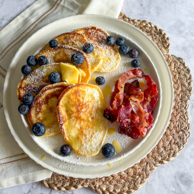 A sweet and savory breakfast plate featuring a stack of diner-style buttermilk pancakes, crispy bacon strips, and fresh blueberries.