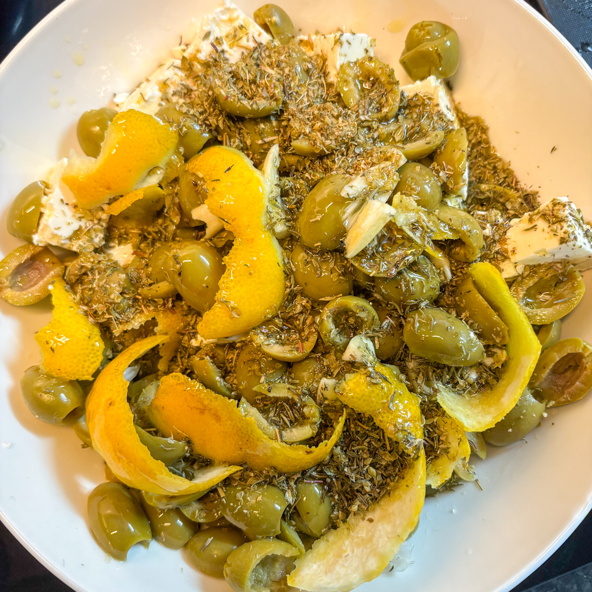 Green olives, feta, lemon peel and herbs ready to marinate in olive oil