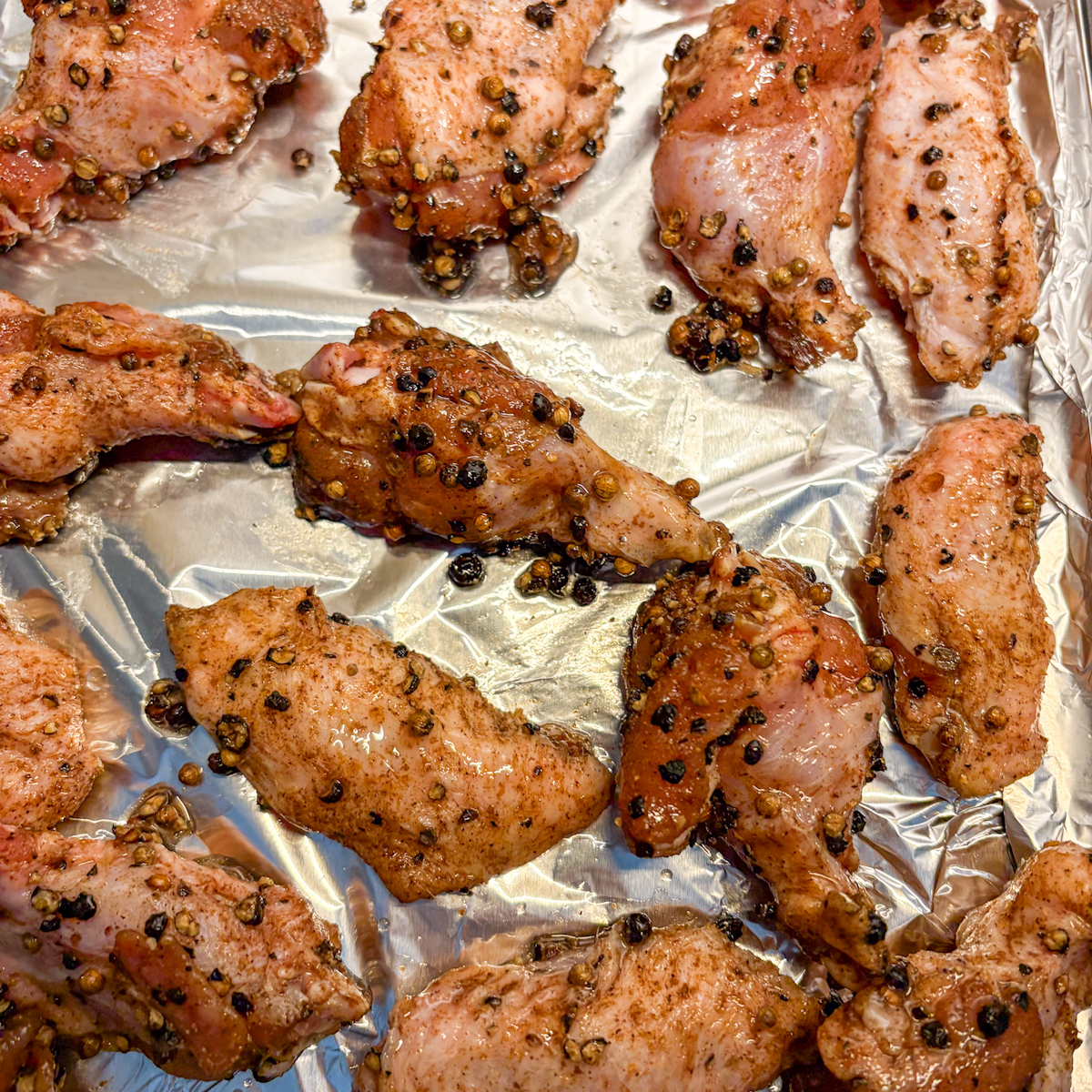Raw chicken wings coated in smashed peppercorn and coriander dry rub on a foil-lined baking tray.