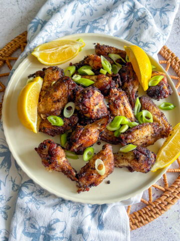 Plate of Crispy Peppercorn Coriander Chicken Wings with lemon wedges and spring onions on a wicker placemat.