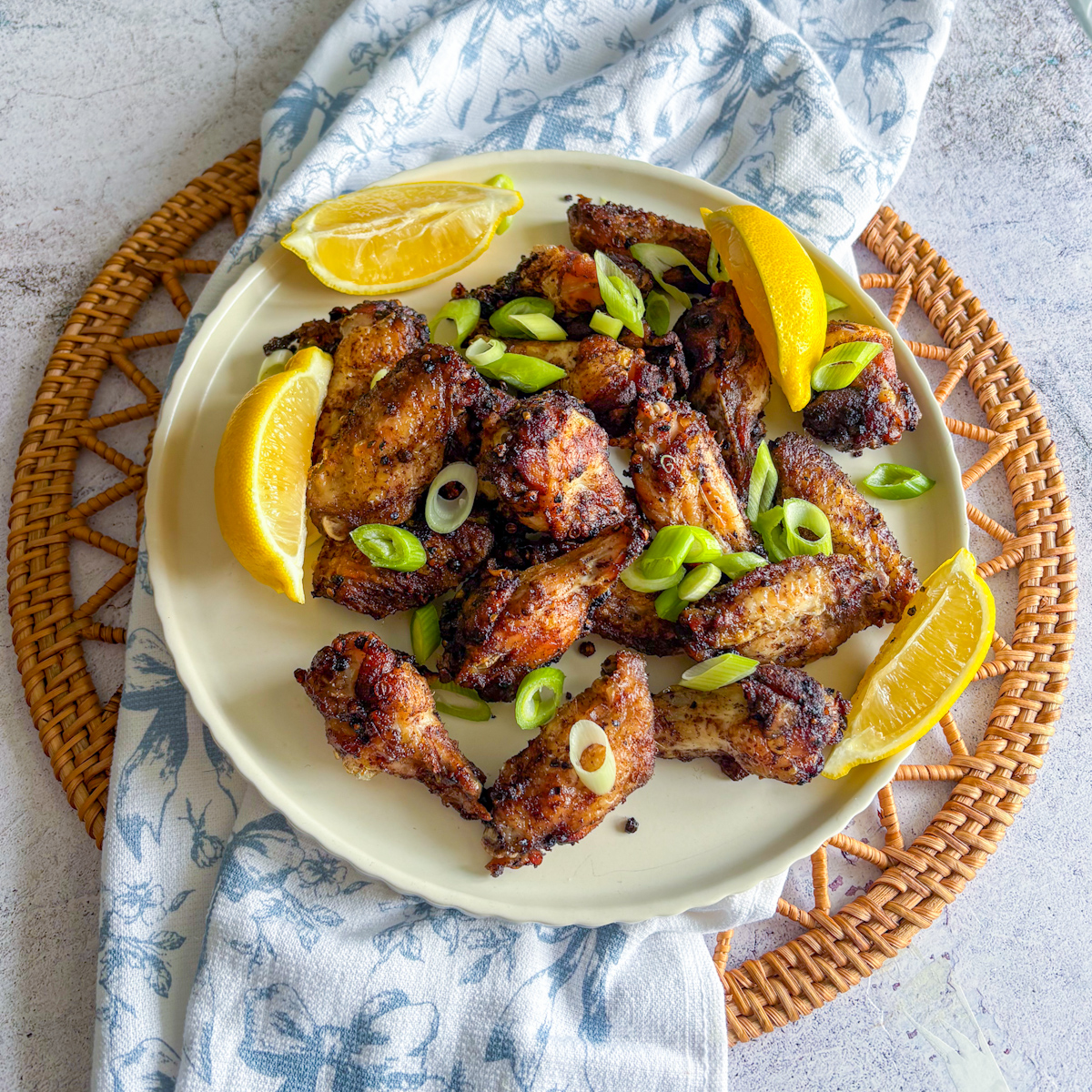 Plate of Crispy Peppercorn Coriander Chicken Wings with lemon wedges and spring onions on a wicker placemat.