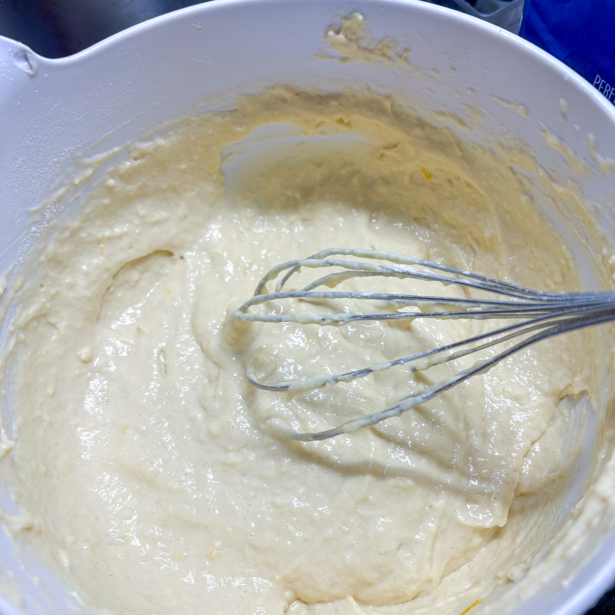 A top-down view of a balloon whisk mixing plain flour into the lemon ricotta base to form a smooth batter for Ricotta Pancakes in a white bowl.