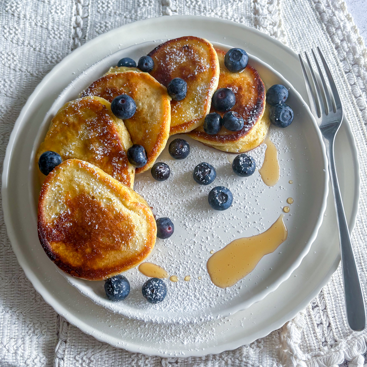 : An overhead view of the best ricotta pancakes recipe served on a white plate with a silver fork on a lace tablecloth.