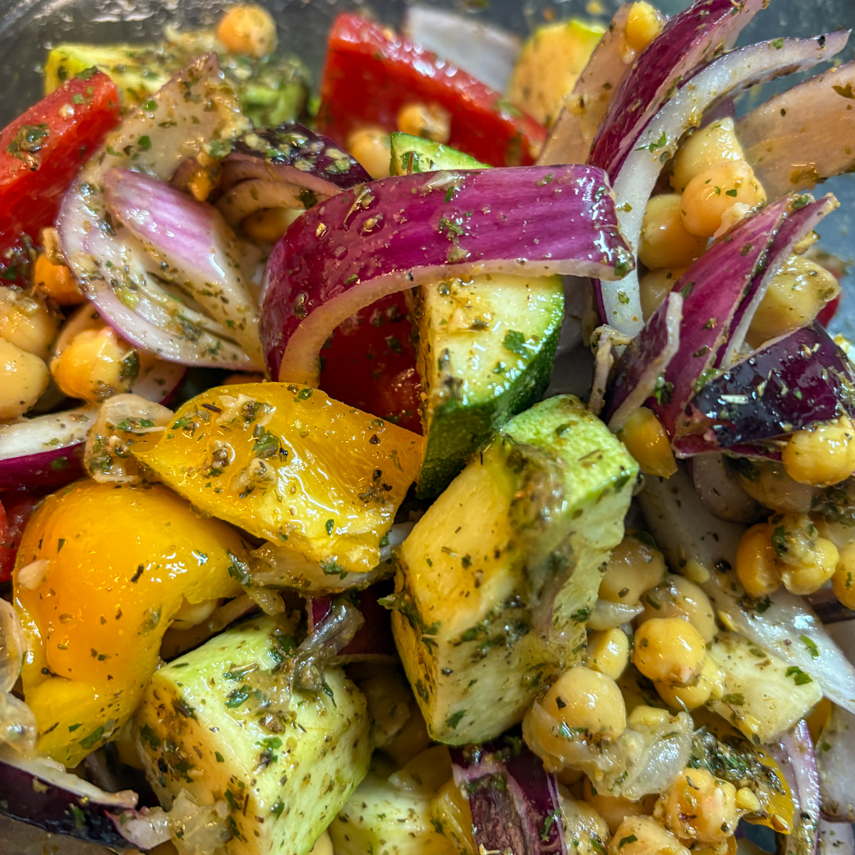 Close-up of raw courgettes, peppers, red onion, and chickpeas tossed in a fragrant herb oil in a bowl, the first step for making roasted vegetables and halloumi.