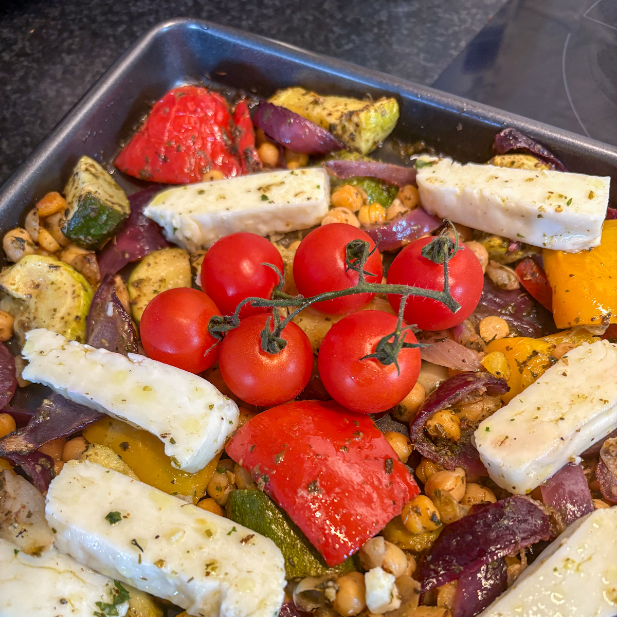 A baking tray filled with partially roasted vegetables and halloumi slices, topped with fresh cherry tomatoes on the vine before the final bake.