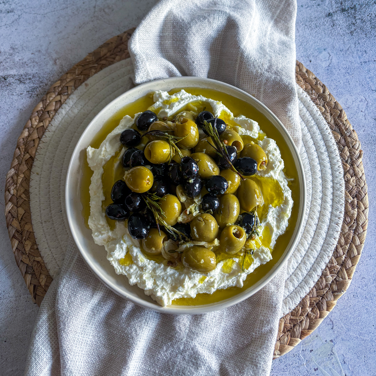 Overhead shot of glossy marinated olives with rosemary sprigs on whipped feta dip