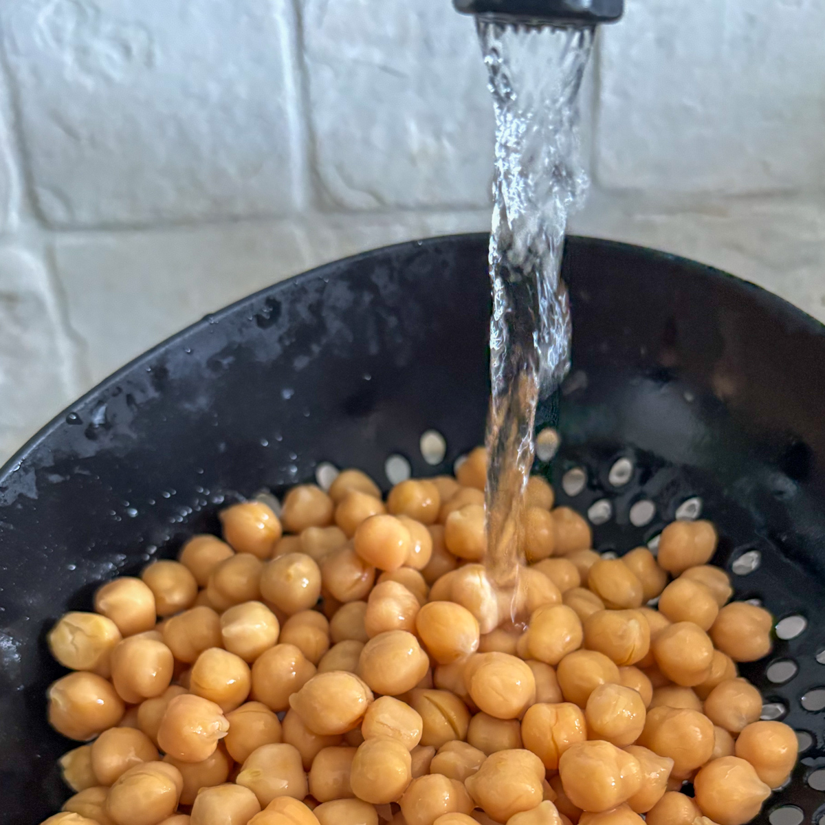 Rinsing tinned chickpeas under running water in a black colander, the first step in how to pan-fry tinned chickpeas.